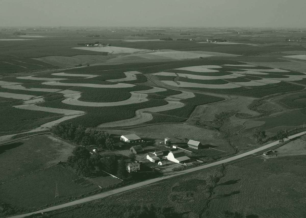 Aerial view of rolling Iowa farmland in curved contour strips, with a farmstead and a straight gravel road running to the horizon