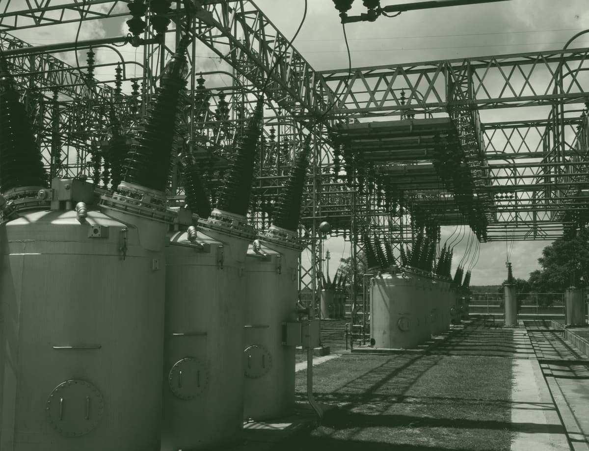 A substation switchyard: rows of oil circuit breakers with tall insulator bushings under a steel lattice frame, on mown grass