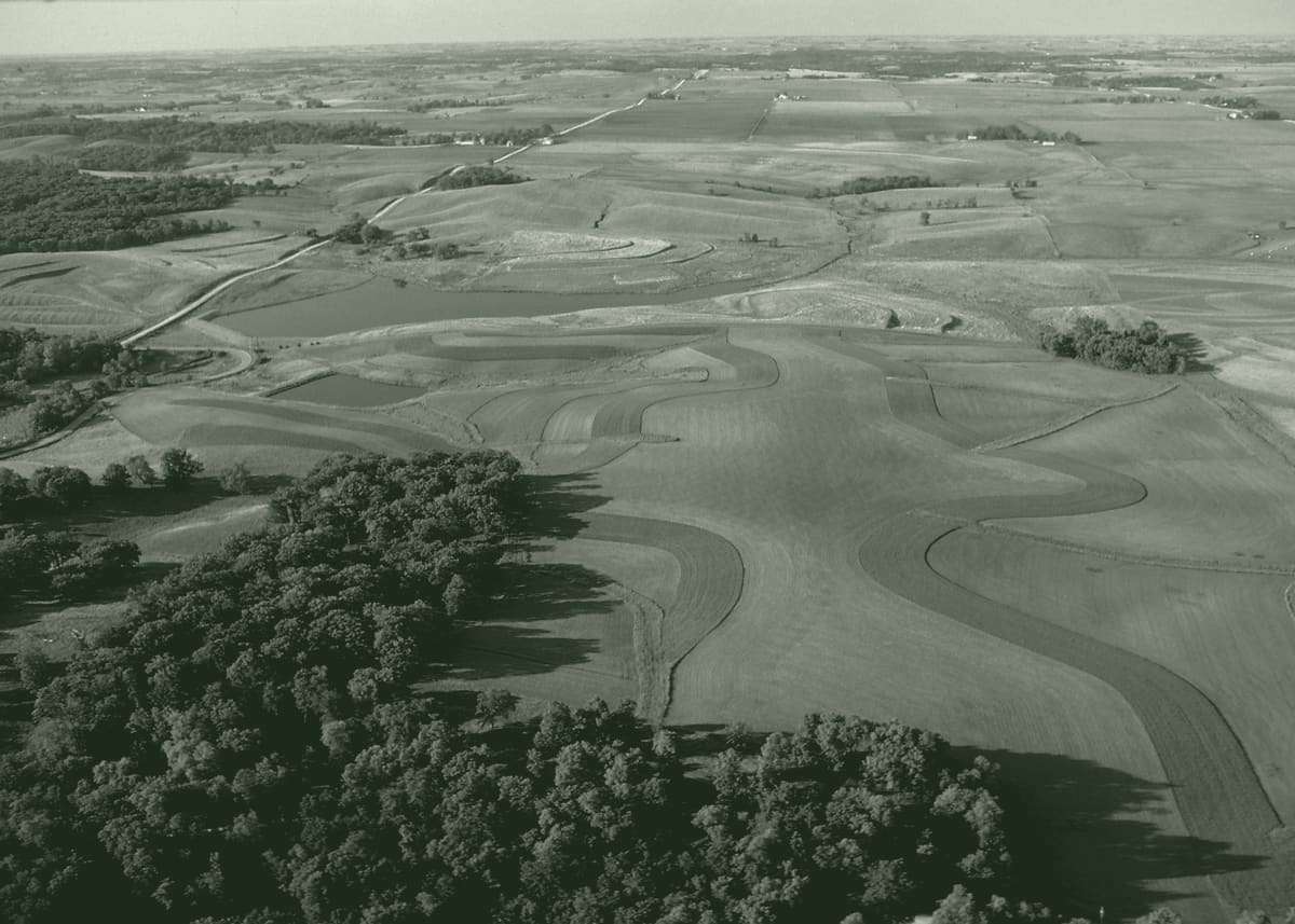 Aerial view of a patchwork of fields, terraces and grass buffer strips around a small lake, with woodland in the foreground